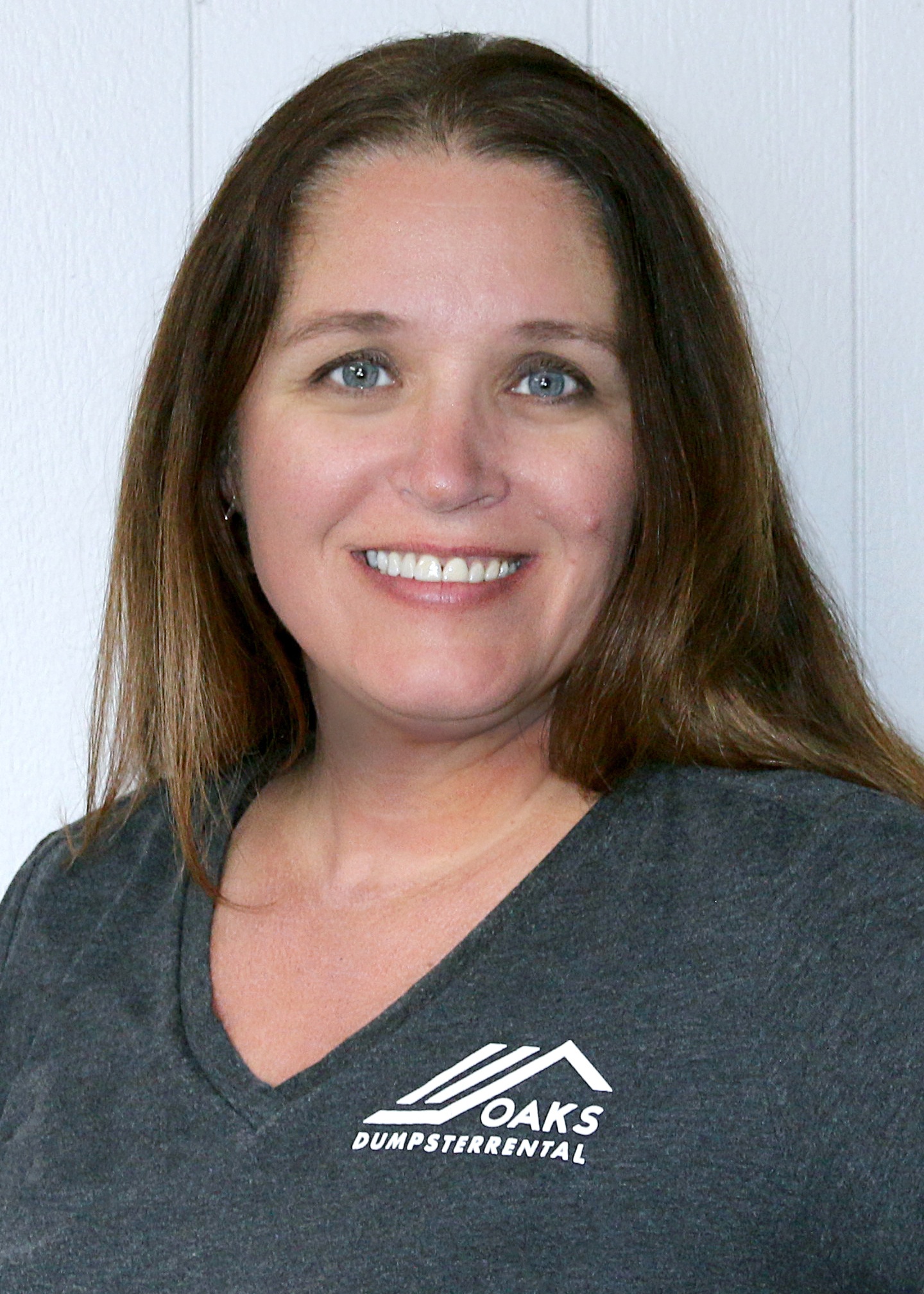 Woman with long brown hair and gray eyes smiling at the camera. She is wearing a dark grey t-shirt with a logo on it.