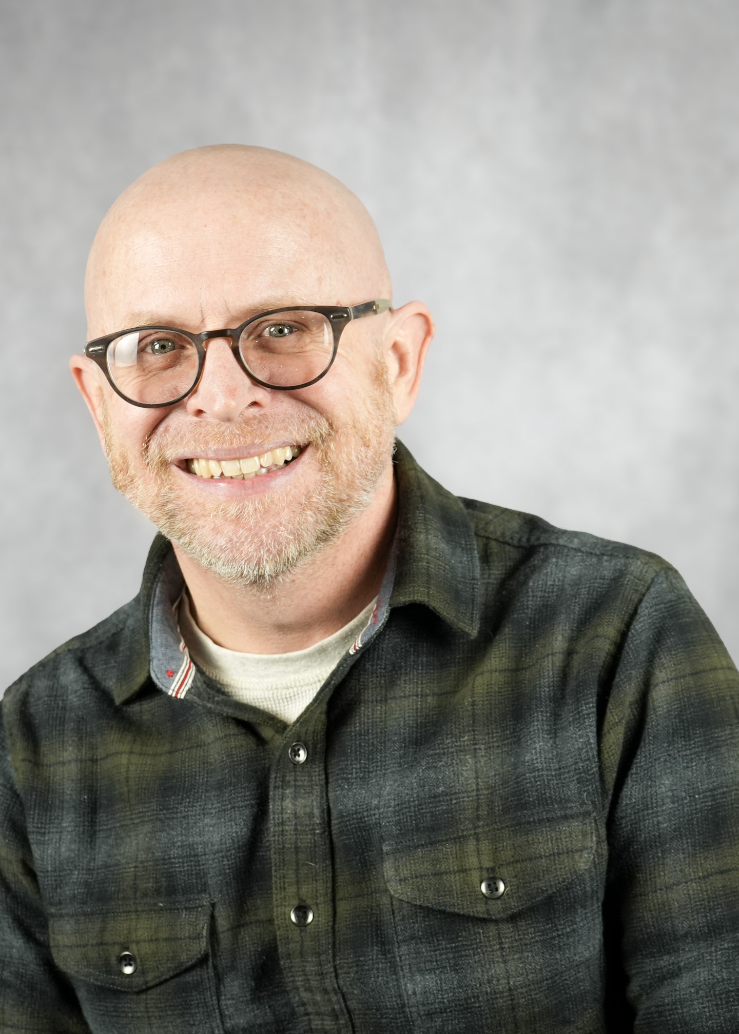 A man with a warm smile wearing glasses and a plaid shirt. He appears to be in a professional setting.