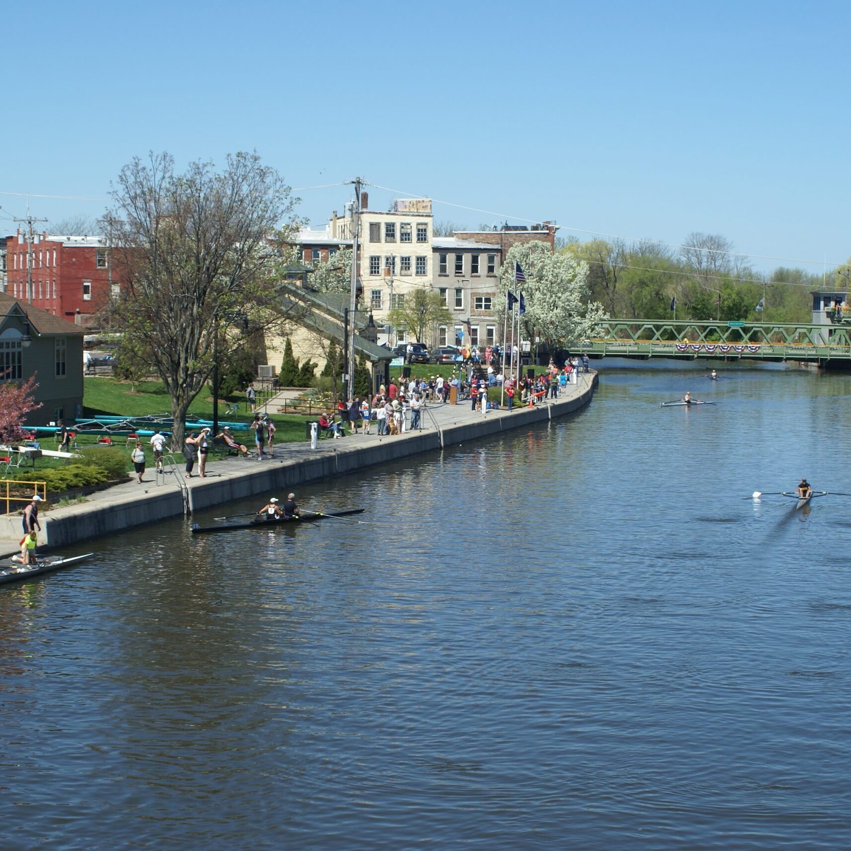 A river with people rowing in a boat and others walking along a path beside it. The scene is set on a sunny day with trees and buildings in the background.