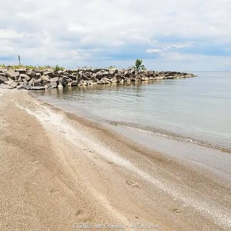 A tranquil beach scene with a rocky shoreline and calm sea. The sandy beach stretches towards a pile of rocks.