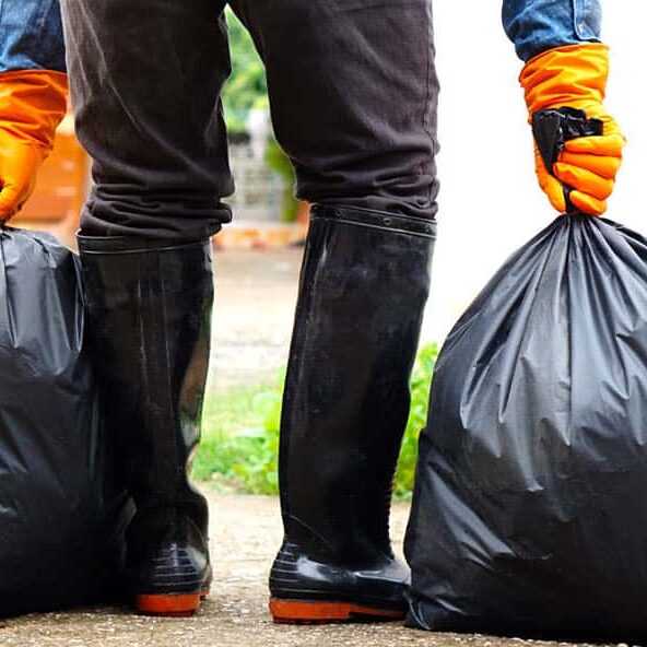 A person wearing orange gloves and black boots holding trash bags.